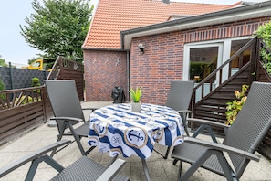 Une terrasse avec une table ronde recouverte d’une nappe aux motifs marins, entourée de chaises en métal, et un bâtiment en briques avec un toit recouvert de tuiles rouges en arrière-plan.