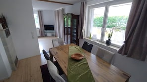Zona pranzo con tavolo in legno, sedie, una televisione sullo sfondo e vista sul verde all'esterno.