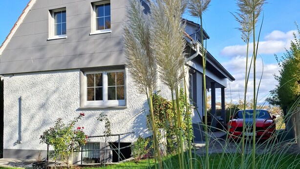 Une maison moderne avec un toit en pente, une petite fenêtre et un garage dans lequel est garée une voiture rouge.