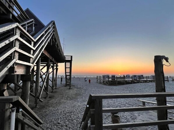 Un muelle de madera con una escalera que lleva a un edificio, con vista a una playa donde hay gente y un atardecer.