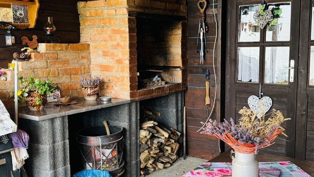 Une cuisine rustique avec un four en briques, des étagères en bois et une table recouverte d’une nappe à motifs floraux.