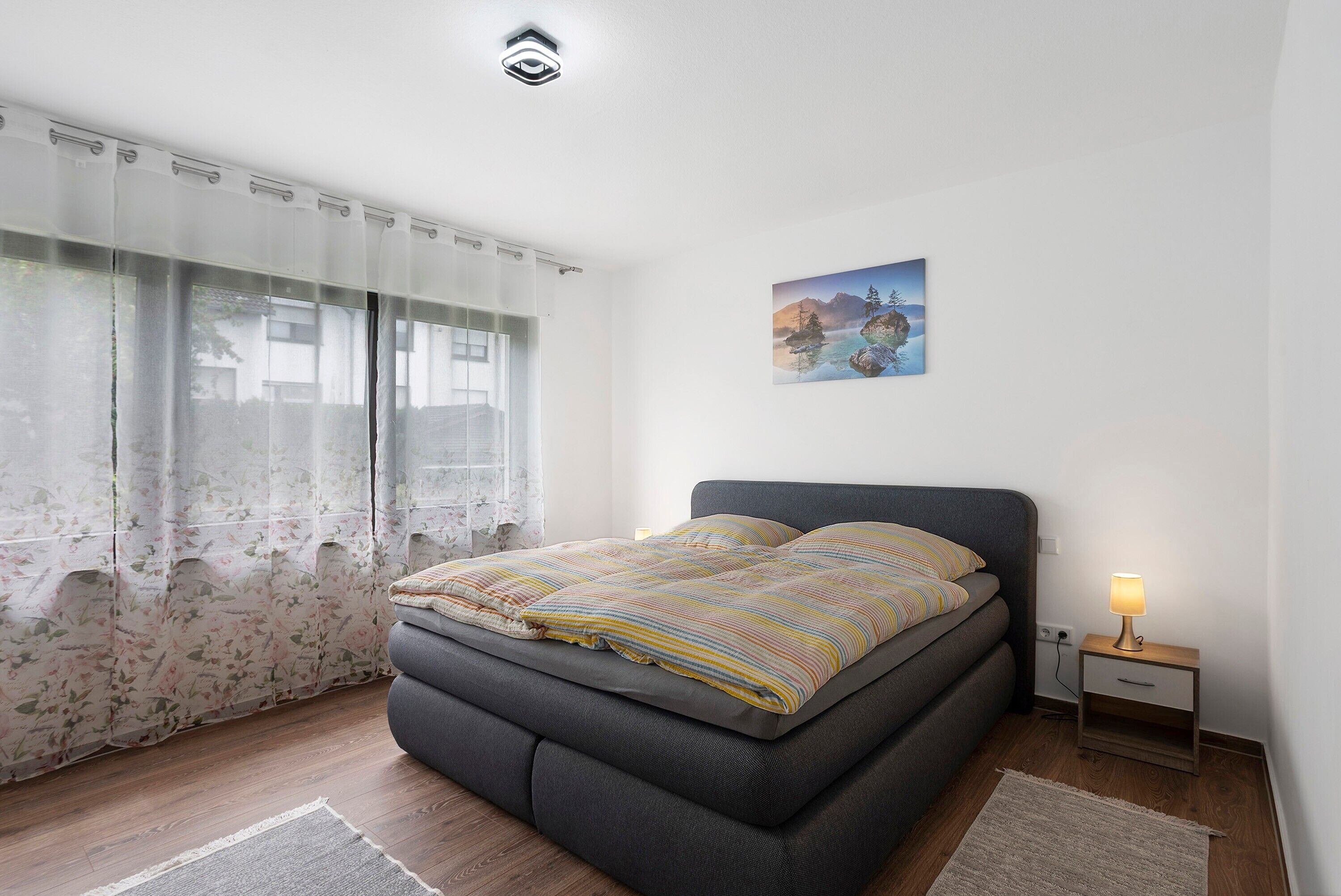 A bedroom with a bed, a nightstand with a lamp, a painting on the wall, and sheer curtains.