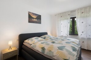 Un dormitorio con cama, mesita de noche, lámpara de pared, ventana con cortinas y un cuadro en la pared.