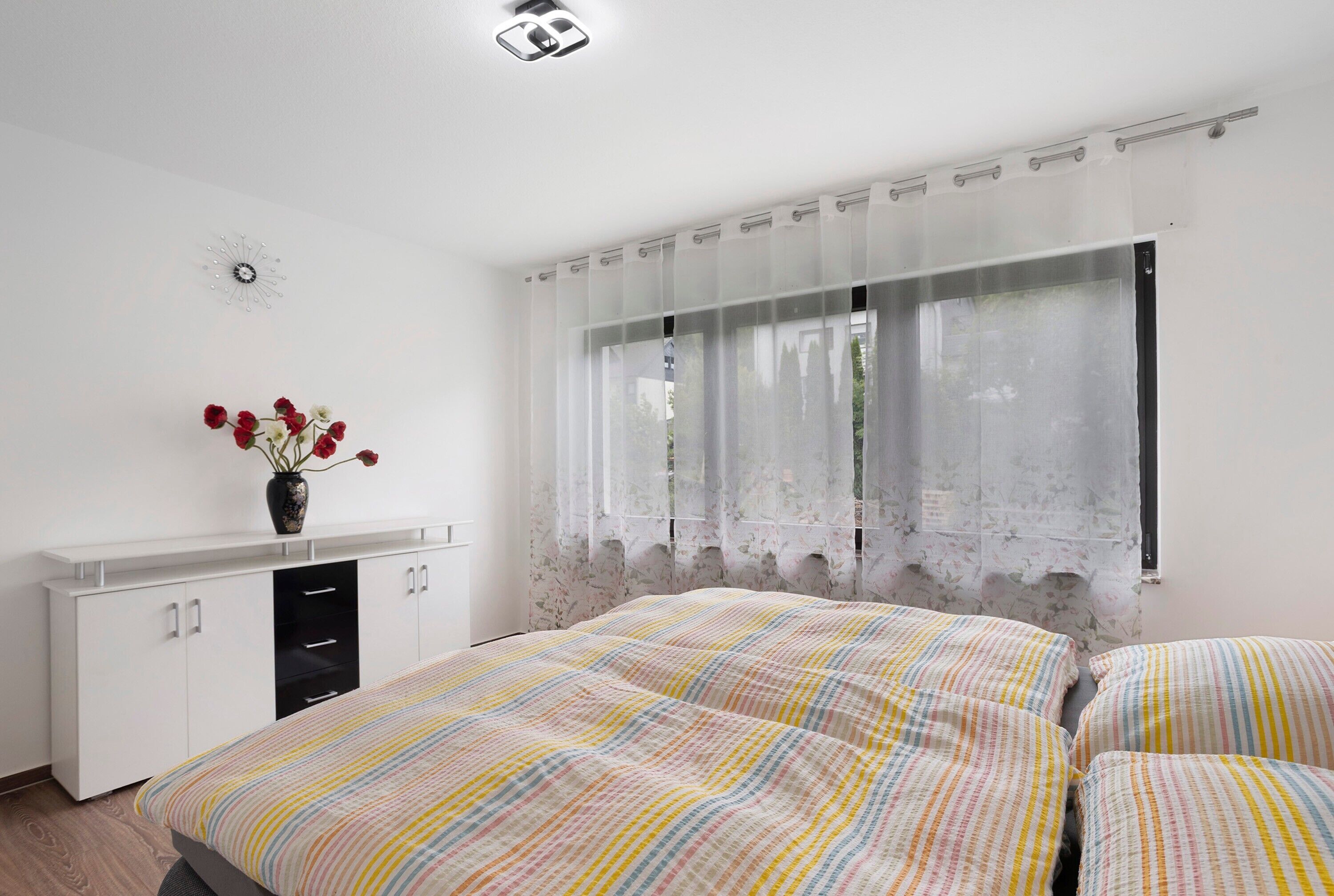 Une chambre à coucher avec un lit, une armoire blanche, une fenêtre avec des rideaux légers et un vase rempli de fleurs.