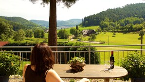 Une femme est assise à une table en bois, devant une bouteille de vin et deux verres. Elle a devant elle un paysage verdoyant, avec des collines et une maison qui se dessine à l'horizon.