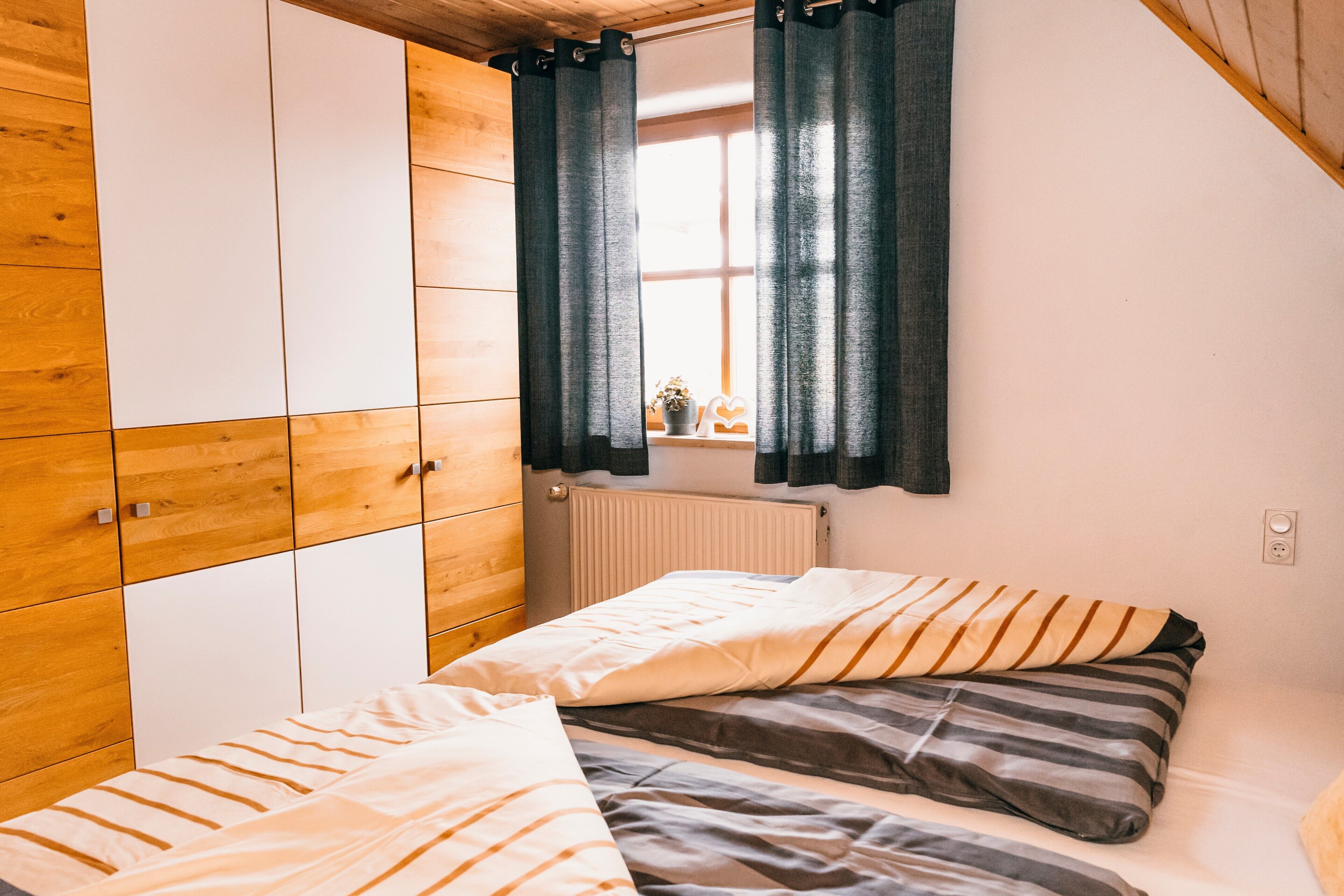 A bedroom with two beds, wooden paneling on the walls, a window with curtains, and a radiator.
