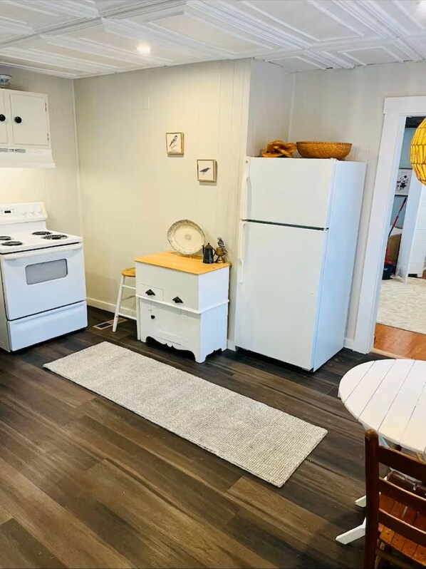 Una cocina compacta con armarios blancos, estufa, refrigerador y una pequeña mesa con sillas.