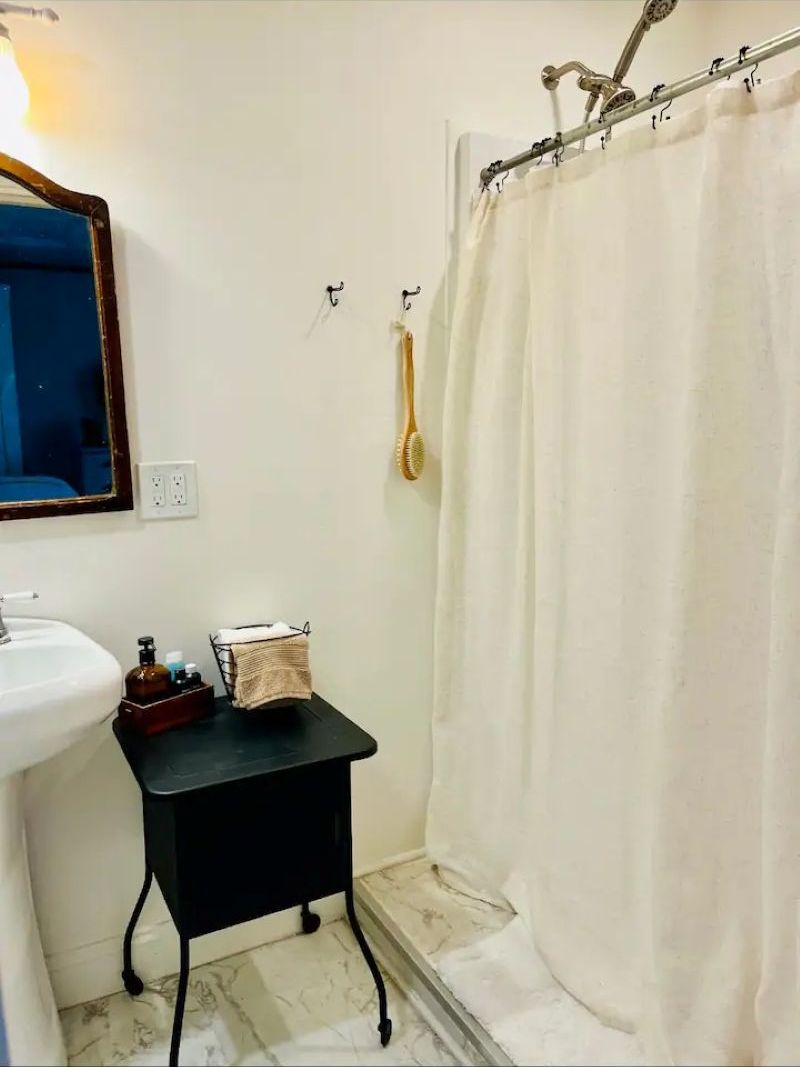 Kylpyhuoneessa on valkoinen pesuallas, peili, musta sivupöytä, jossa on hygieniatuotteita, ja valkoinen suihkuverho.