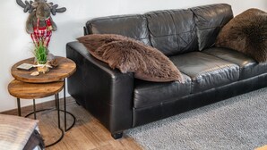 Un canapé en cuir noir agrémenté d’un plaid en fausse fourrure marron, une petite table en bois avec une plante, et une décoration représentant un élan accrochée au mur.