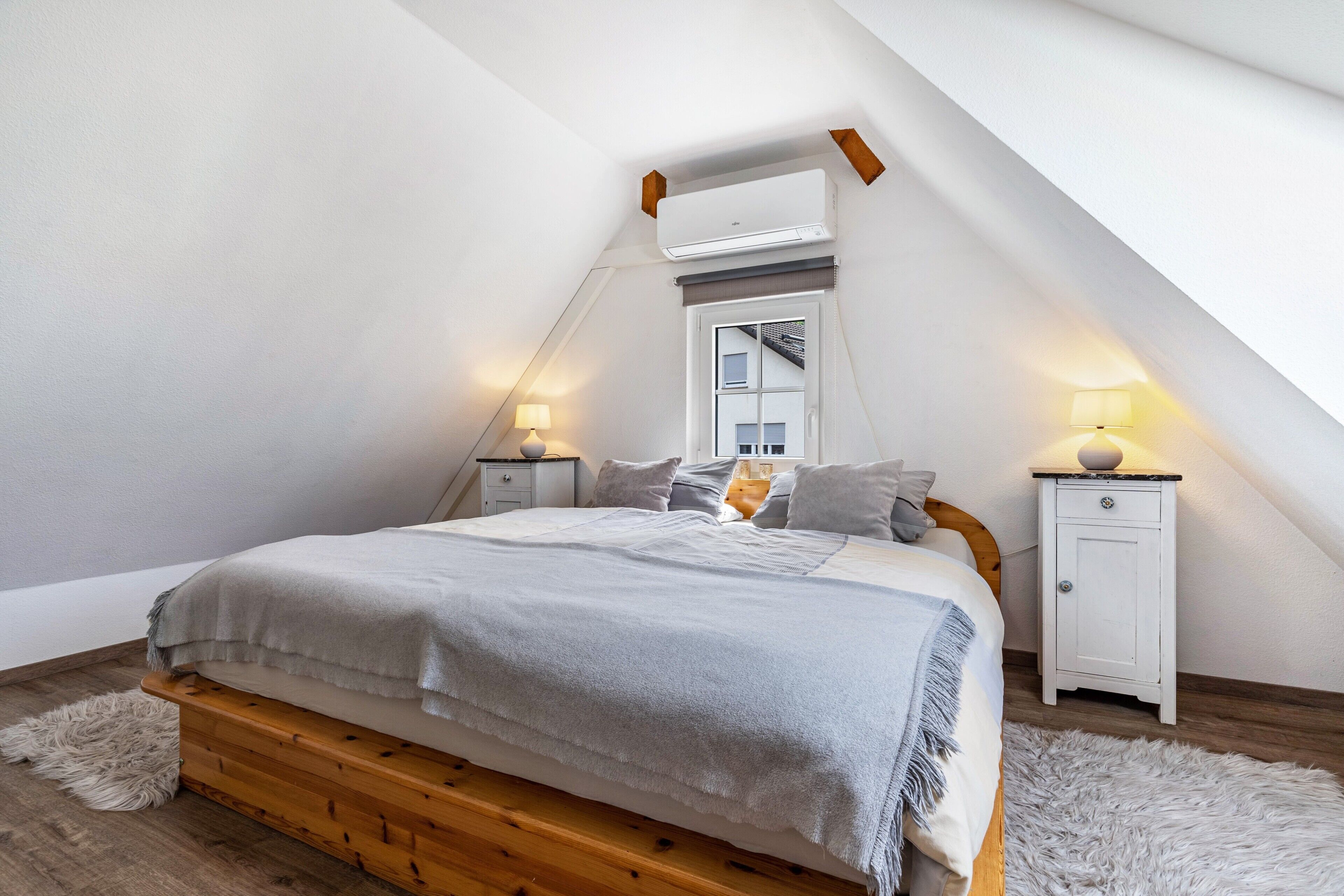 Ein Schlafzimmer mit schrägem Dach, einem Bett mit Holzrahmen, einem weißen Nachttisch, einer Lampe und einem flauschigen Teppich.
