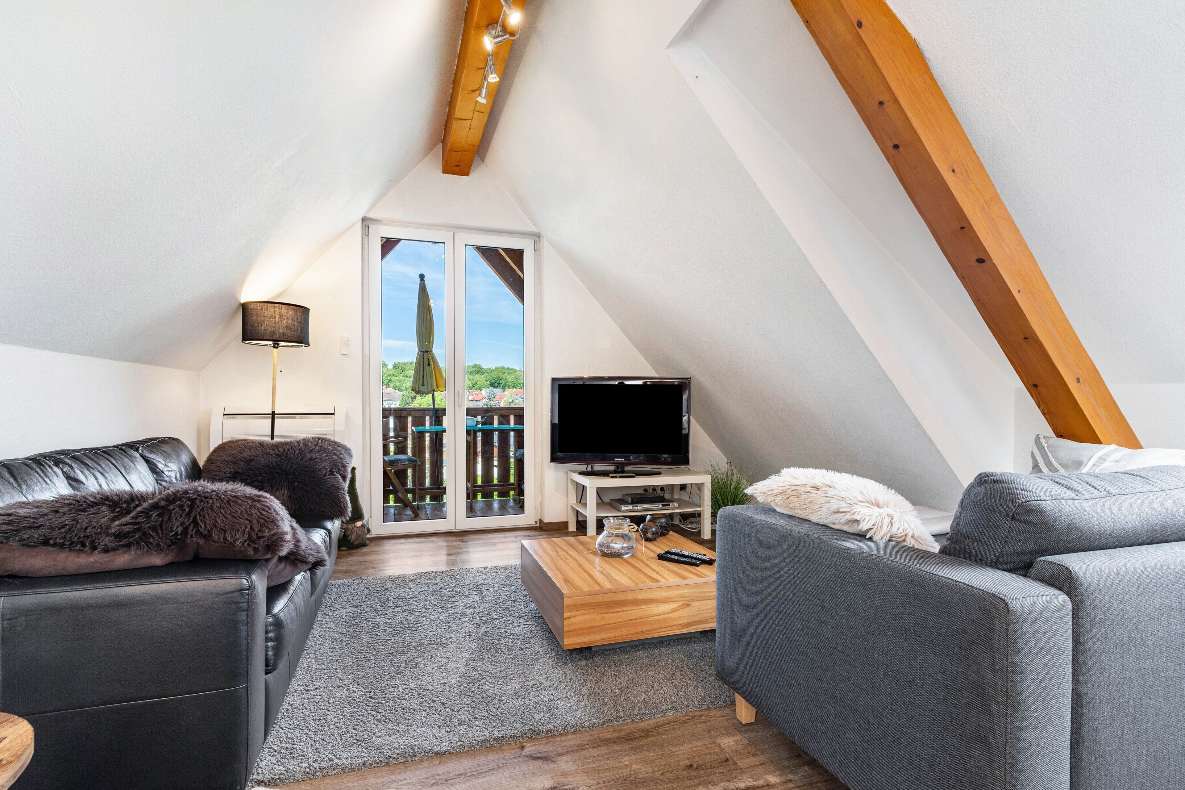 Ein modernes Schlafzimmer im Loft-Stil mit schrägverdeckter Decke, großem Fenster, Flachbildfernseher und einer gemütlichen Sitzecke mit Sofa und Sessel.