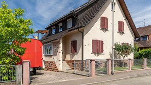 Une maison à deux étages avec un réservoir rouge à l’extérieur, une allée fermée par un portail et un ciel dégagé.
