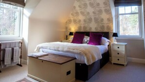 A bedroom with a sloped ceiling, a bed with white bedding and purple pillows, a bedside table with a lamp, and a window with a view of trees.