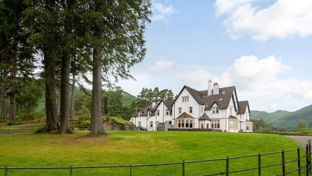 A large, white, multi-story house with a prominent chimney, surrounded by lush green fields and trees.
