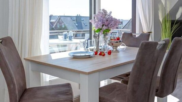 A dining area with a table set for four, a vase with flowers, and a view of buildings through large windows. - Haus Belvedere - Directly behind the dune crest, only 50m to the beach crossing - (Sylt)