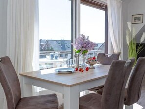 A dining area with a table set for four, a vase with flowers, and a view of buildings through large windows. - Haus Belvedere - Directly behind the dune crest, only 50m to the beach crossing - (Sylt)
