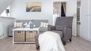 A modern living room with a grey armchair, a grey sofa, and a white coffee table. There are wicker baskets on the coffee table and a white blanket with fish patterns on the floor. - Haus Belvedere - Directly behind the dune crest, only 50m to the beach crossing - (Sylt)