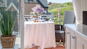 A balcony with a table set for two, a wicker plant, and a view of a house and hills. - Sea view, House Belvedere, directly behind the dune crest, for up to 5 people - (Sylt)