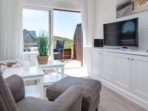 A modern living room with a sofa, a coffee table, and a TV on a cabinet. There is a view of a balcony with a plant and a chair. - Sea view, House Belvedere, directly behind the dune crest, for up to 5 people - (Sylt)