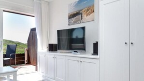 A modern living room with a flat-screen TV, white cabinets, a grey ottoman, and a view of a balcony with outdoor furniture. - Sea view, House Belvedere, directly behind the dune crest, for up to 5 people - (Sylt)