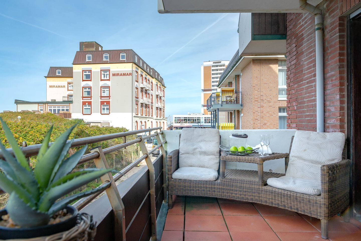 Un balcon agrémenté de meubles en osier, d’une plante en pot et offrant une vue sur les bâtiments qui se dressent à l’arrière-plan.