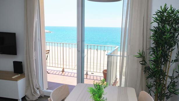 A room with a balcony overlooking the sea, a dining table with chairs, a TV on a stand, and a potted plant. - Premia Palm View (Premià de Mar)