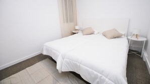 A white bedroom with a bed, two bedside tables, a window with sheer curtains, and a small round table with a lamp. - Premia Palm View (Premià de Mar)