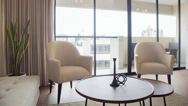 A modern living room with two armchairs, a round coffee table, and a large window with a city view. - w* | Modern 3BR w/ Balcony Near Kennedy Park (Miraflores)
