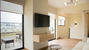 A modern living room with a flat-screen TV mounted on the wall, a sofa, a small table, and a balcony with a view of the city. - w* | Modern 3BR w/ Balcony Near Kennedy Park (Miraflores)