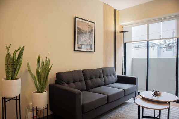 Una sala de estar moderna con un sofá gris oscuro, dos plantas en macetas, un cuadro enmarcado en la pared y una mesa redonda con un objeto decorativo.