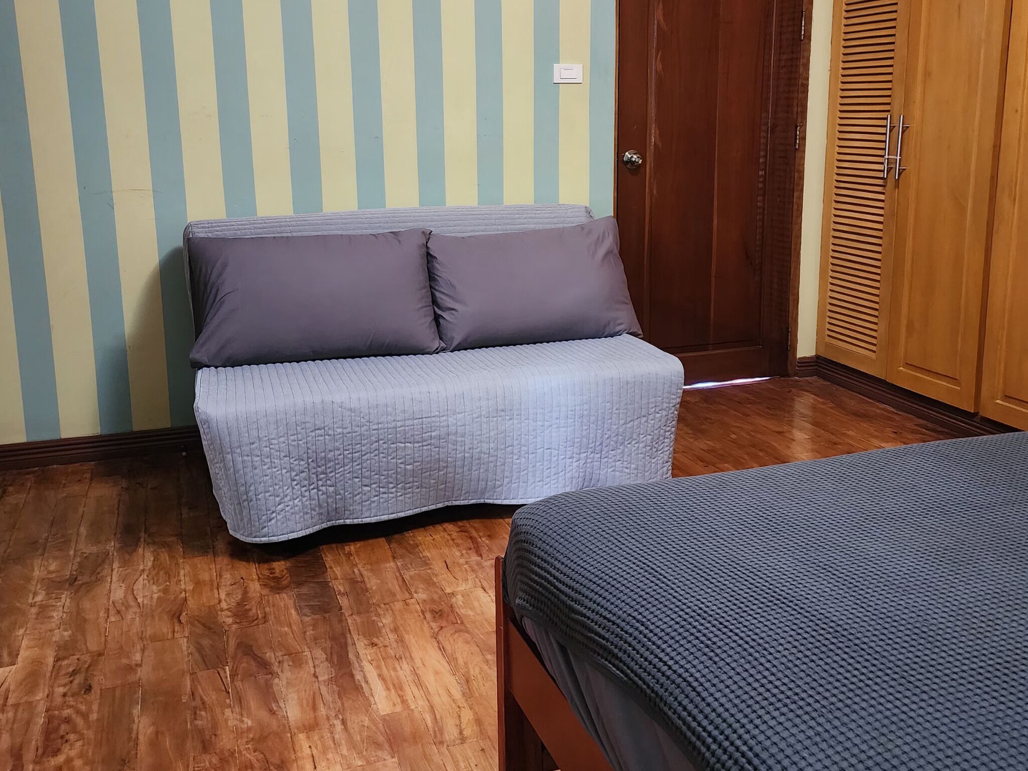 A room with a wooden floor, a sofa with a quilted cover, and a bed with a textured cover.