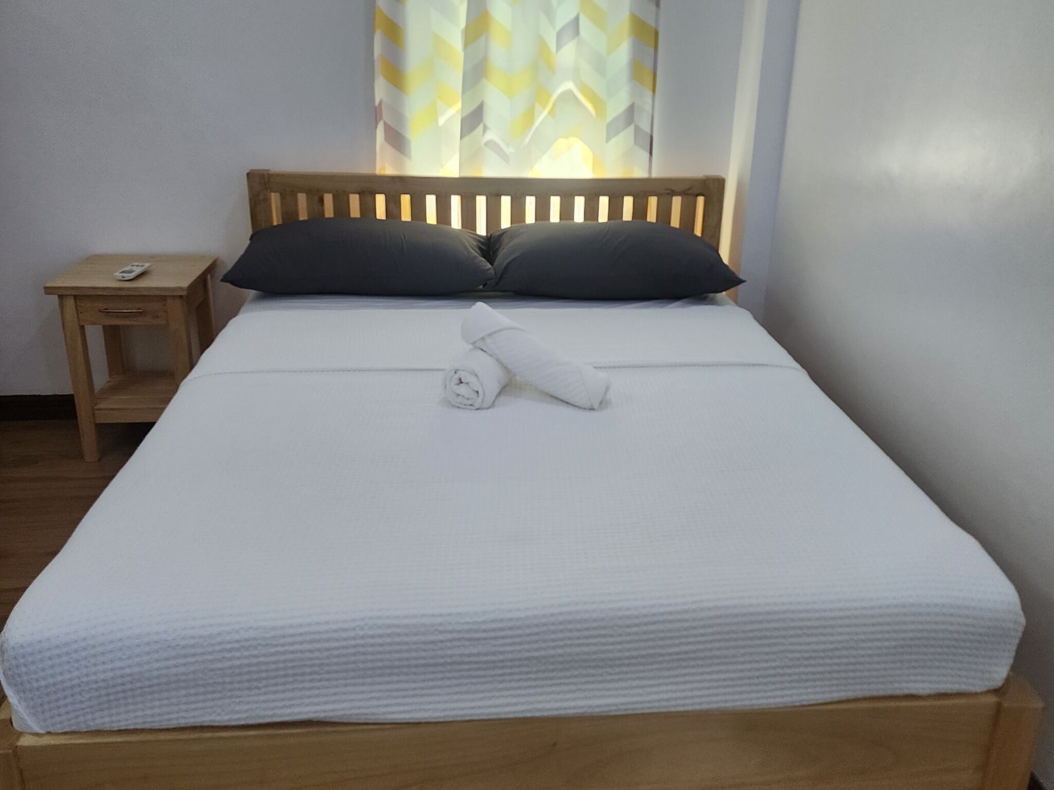 A neatly made bed with white linens, two dark pillows, and a rolled towel.