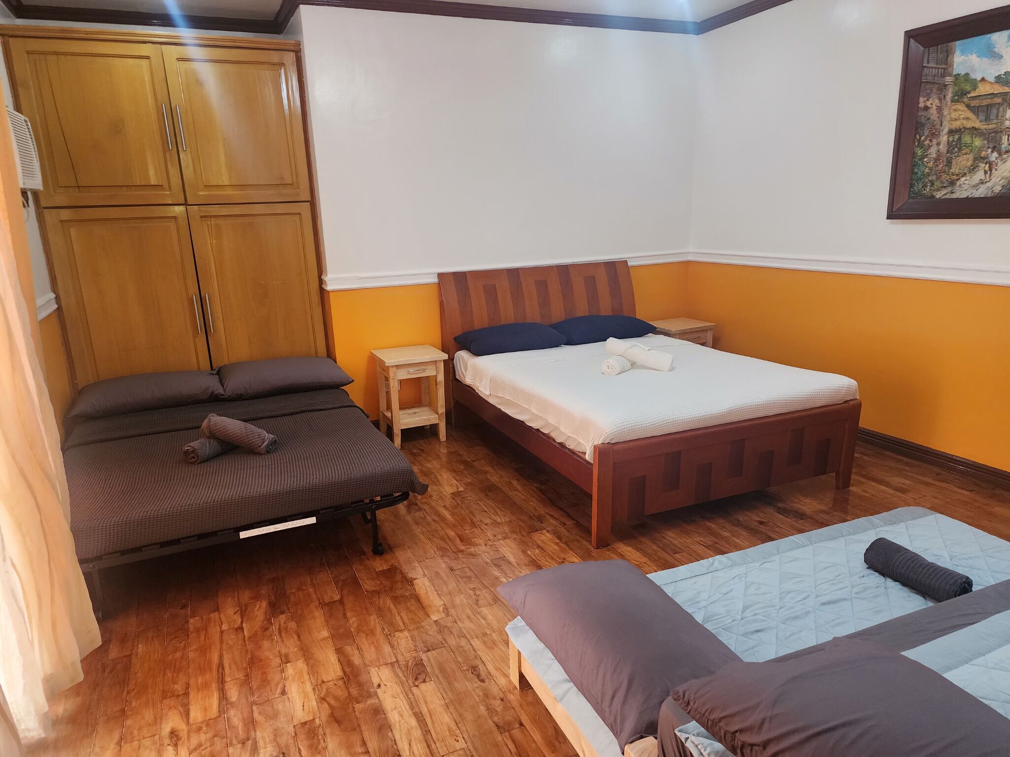 A bedroom with two beds, a wooden wardrobe, a small bedside table, and a painting on the wall.