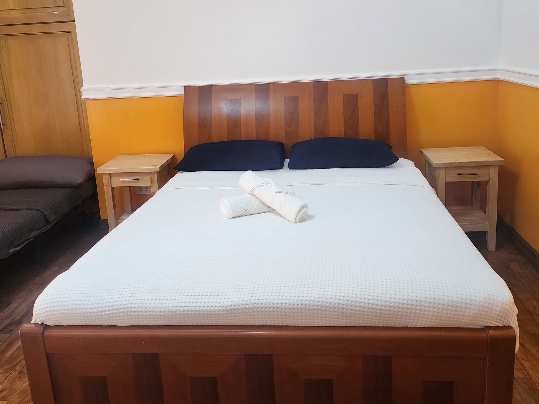 A neatly made bed with a white sheet and two pillows, a wooden headboard, two bedside tables, and a wooden wardrobe.