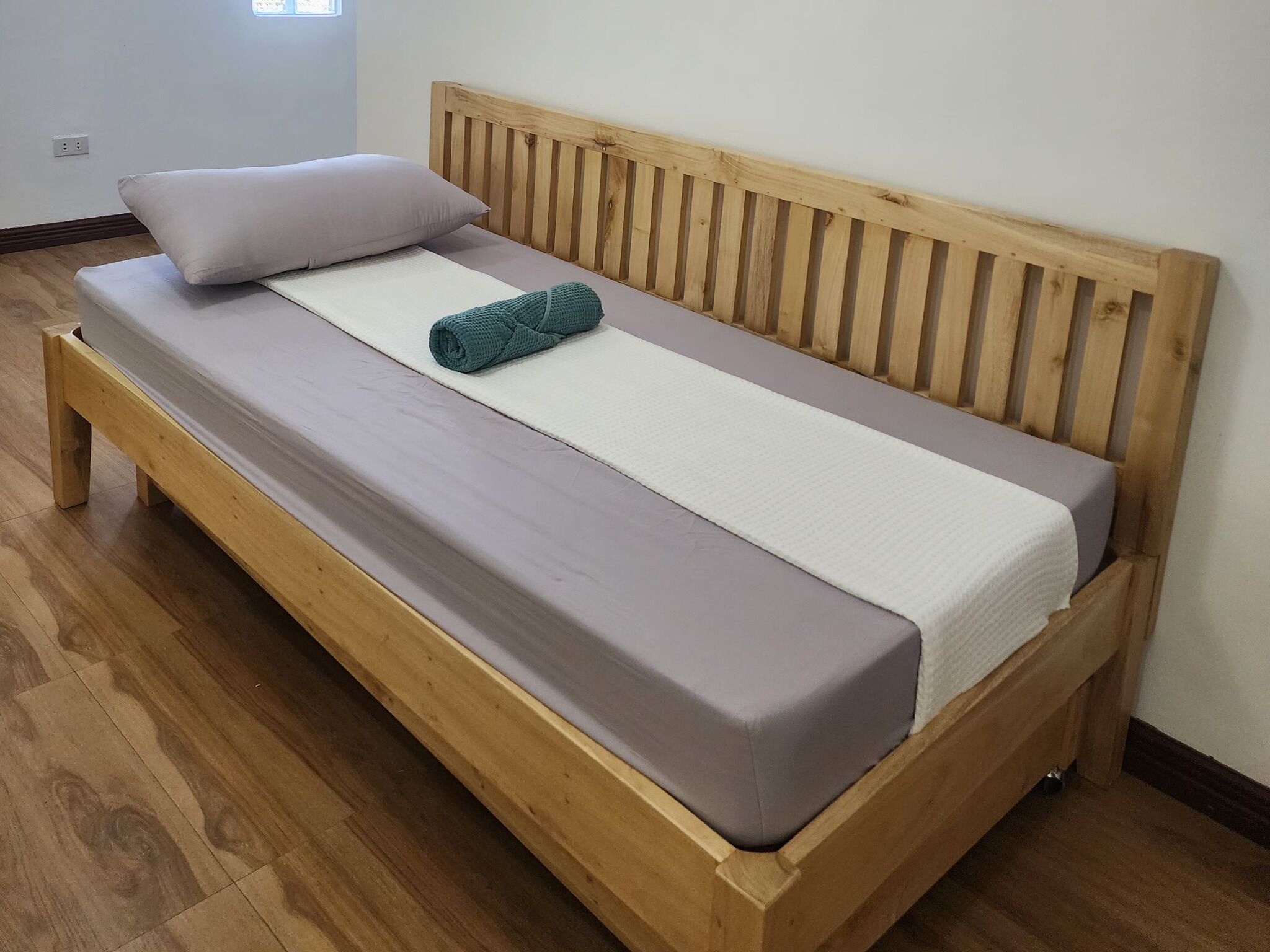 Un letto di legno con testiera, un materasso grigio, un lenzuolo bianco e un asciugamano color petrolio.