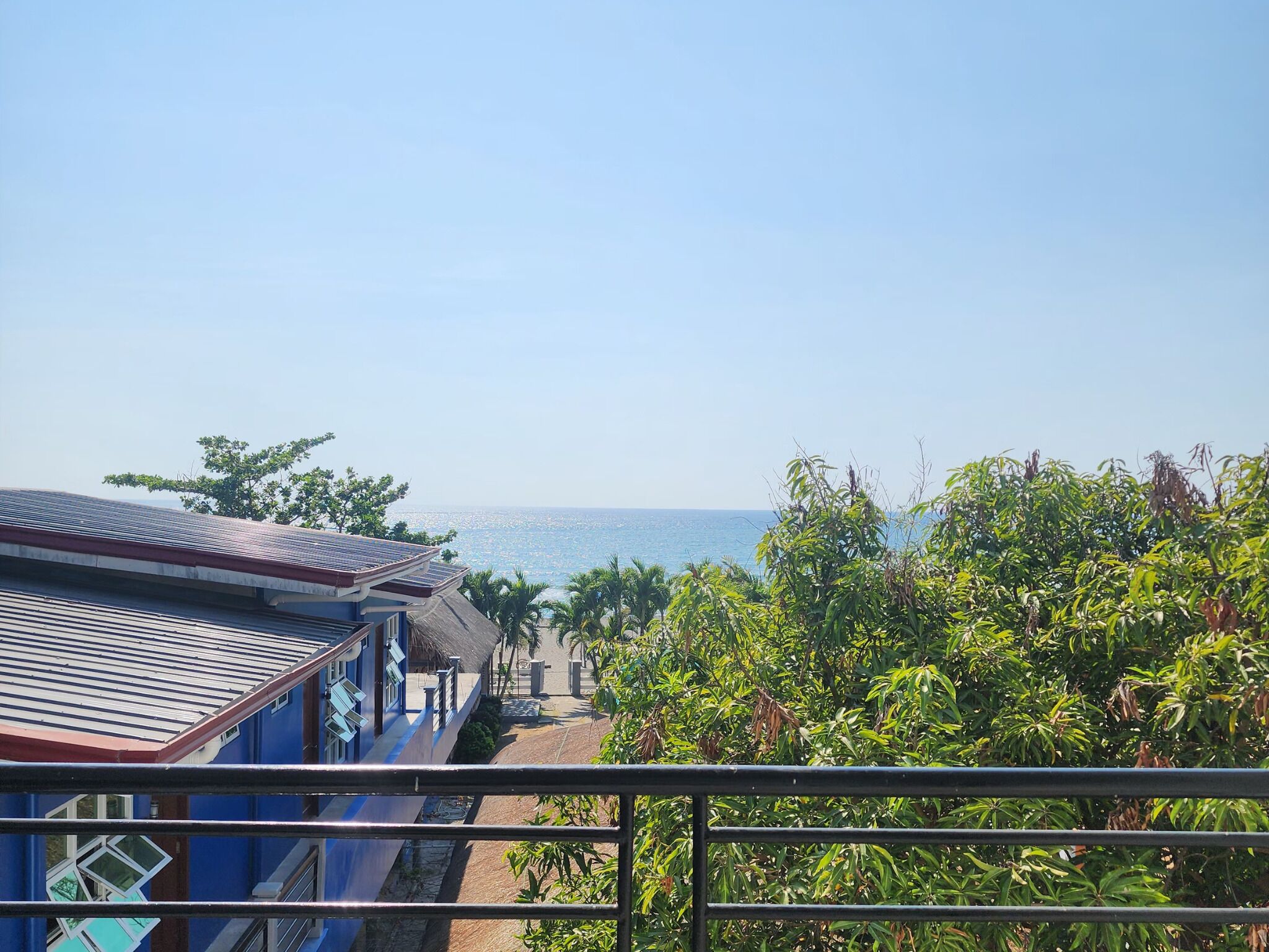 Un hôtel en bord de mer offrant une vue sur l’océan, doté d’un balcon avec une rambarde en métal et d’une végétation luxuriante au premier plan.