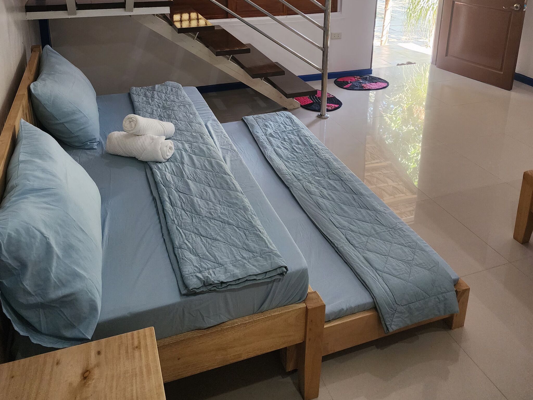 Ein Zimmer mit zwei hölzernen Betten, jeweils mit blauer Matratze und Kissen. In der Ecke steht eine Holzbank und im Hintergrund ist ein Metallgeländer zu sehen.