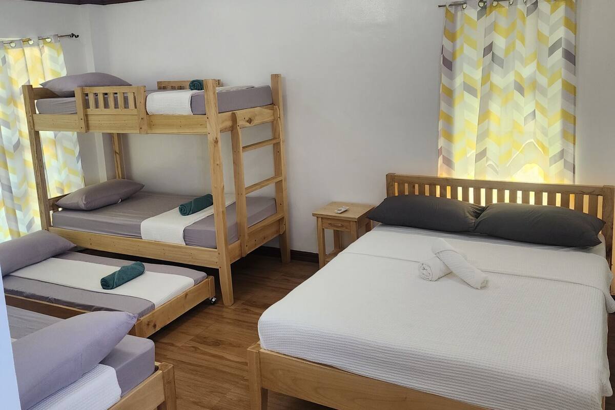 Quarto com duas beliches, uma cama individual, mobiliário de madeira, uma mesinha de cabeceira e cortina com padrão geométrico.