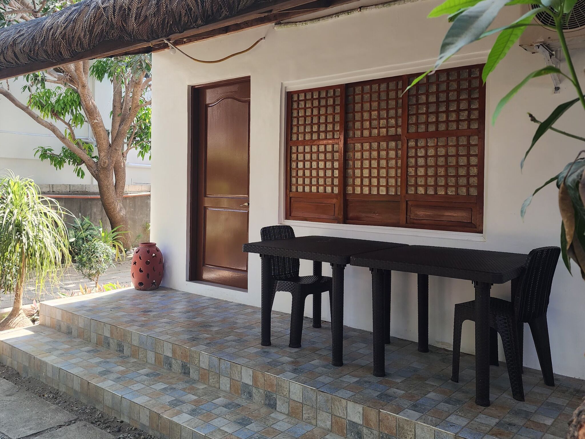 Un patio con piso de baldosas, una mesa con sillas, una puerta grande de madera y una ventana con diseño de cuadrícula.