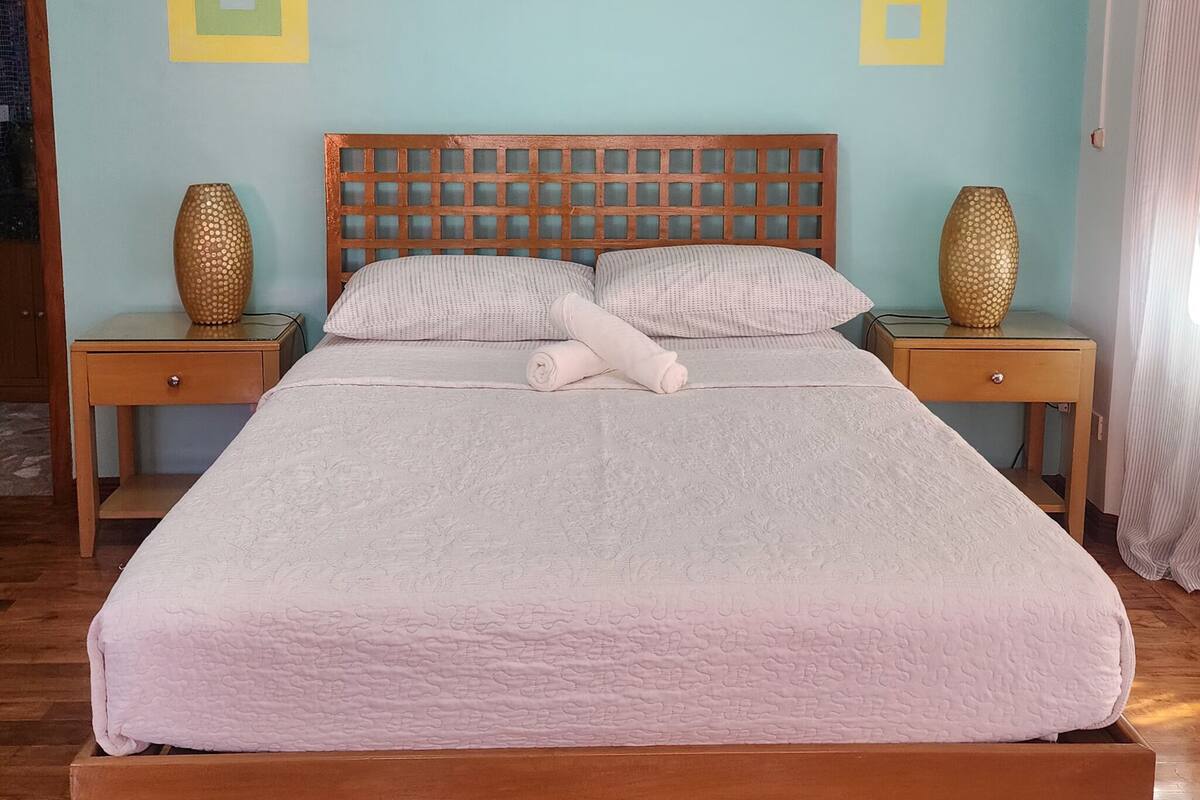 Uma cama bem arrumada com cabeceira de madeira, duas mesas de cabeceira com vasos decorativos e uma parede azul clara com duas decorações quadradas.