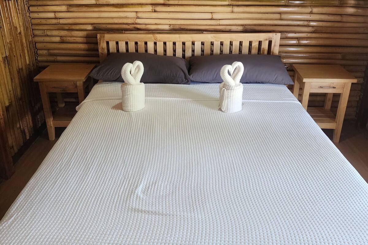 Cama bem arrumada com um espalhadeira branca texturizada, duas toalhas em formato de coração e cabeceira de madeira.
