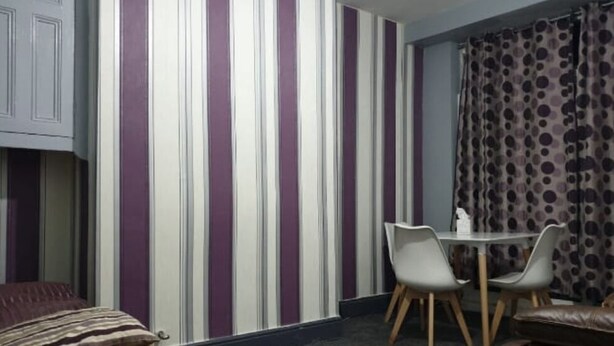 A room with striped wallpaper, a dining table with chairs, a bed, and a window with curtains.