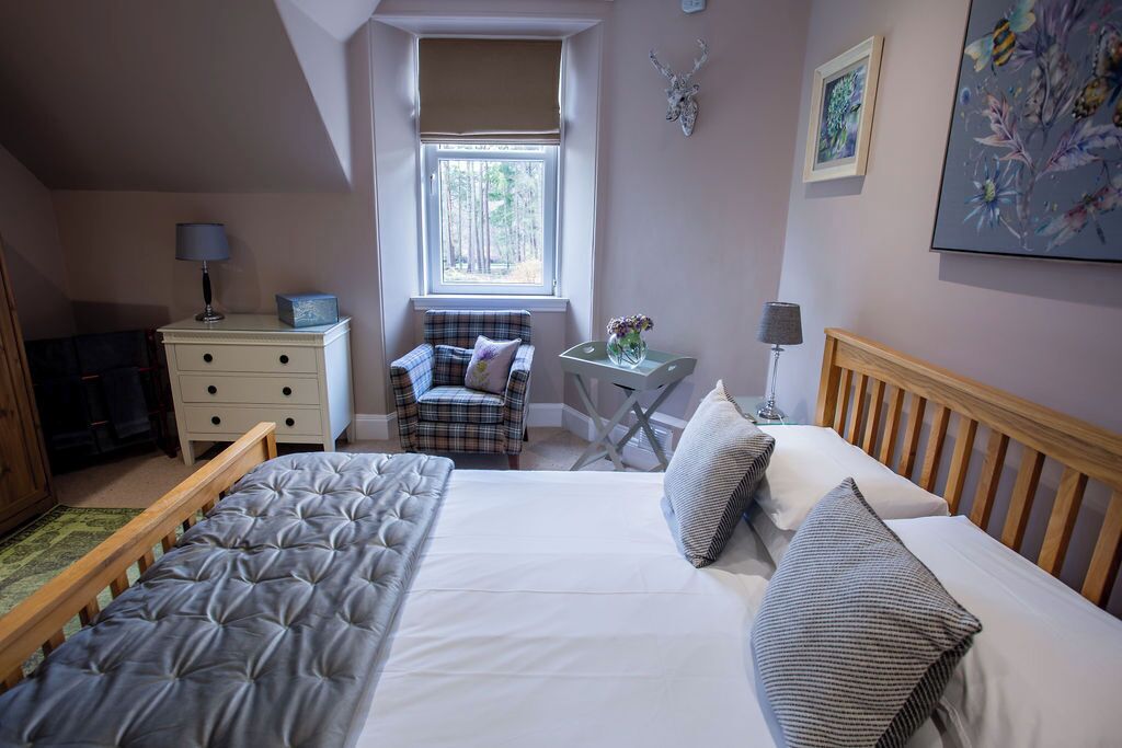 Een slaapkamer met een bed, een stoel, een klein tafeltje met een vaas, een dressoir en een raam met uitzicht op bomen.