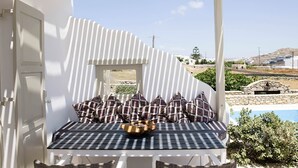 A white room with a checkered table and patterned cushions, a stone wall, and a view of a pool and landscape. - My Rental Homes - Marvi House with shared pool (Mykonos)