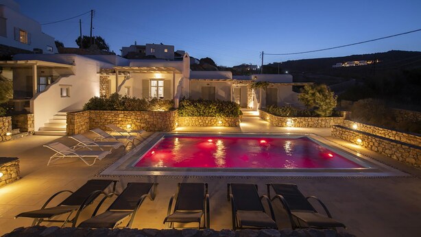 A poolside area with lounge chairs and a swimming pool, surrounded by stone walls and a white building with multiple windows. - My Rental Homes - Marvi House with shared pool (Mykonos)