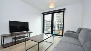 Ein modernes Wohnzimmer mit Flachbildfernseher, Couchtisch aus Holz, grauem Sofa und großem Fenster mit Blick auf Gebäude.