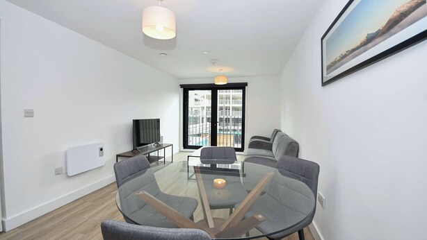 Ein modernes Wohnzimmer mit Glastisch, grauen Sofas, Fernseher und einem großen Fenster mit Blick auf Gebäude.