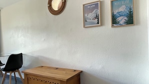Une pièce comprenant un coffre en bois, deux tableaux encadrés représentant des montagnes, un miroir rond et une chaise noire.