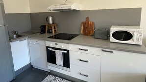 Une cuisine compacte équipée d’un four encastré, d’un four à micro-ondes, d’un grille-pain et d’une machine à café, le tout posé sur un petit tabouret en bois.