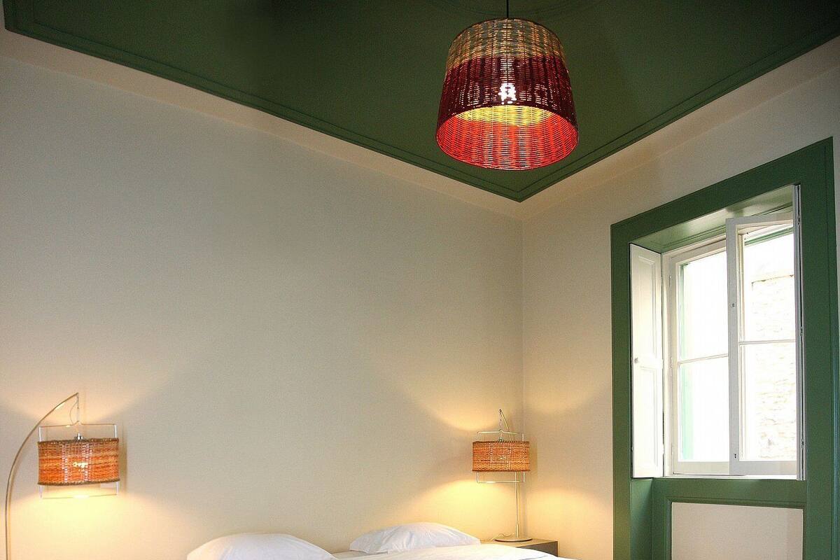 A bedroom with a large bed, two bedside lamps, a window with green shutters, and a hanging red pendant light.